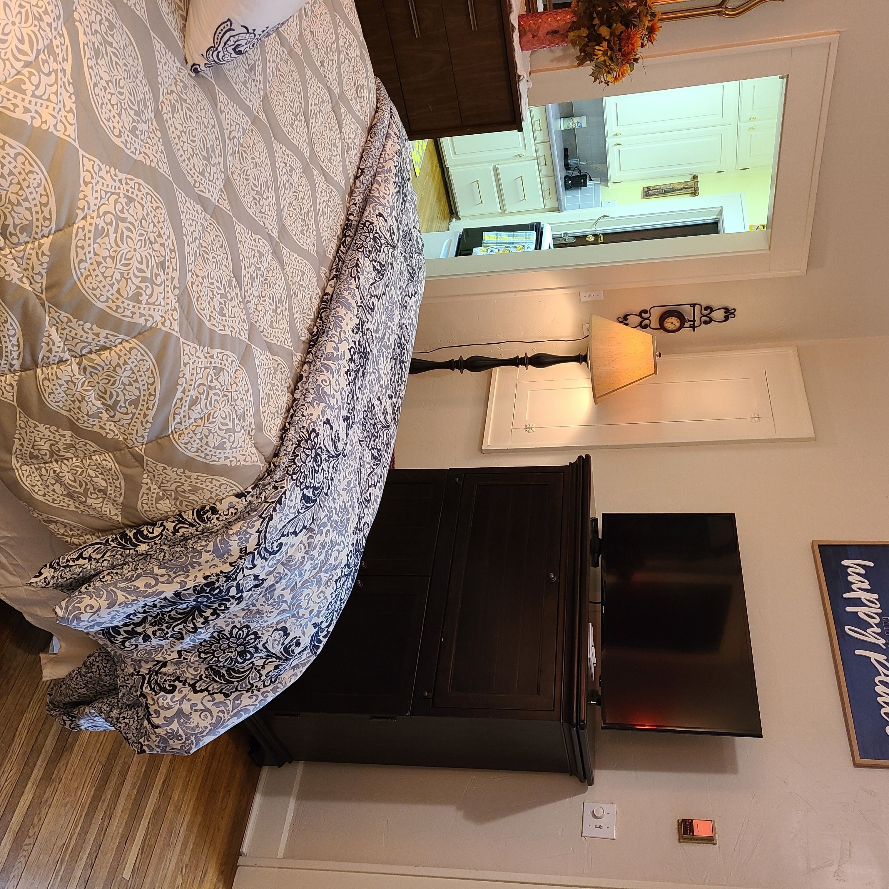 Un dormitorio con una cama, un televisor sobre un aparador, una lámpara y una cocina al fondo.