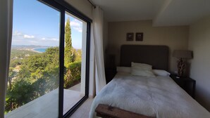 Un dormitorio con un ventanal grande, una cama, una mesita de noche y una lámpara.