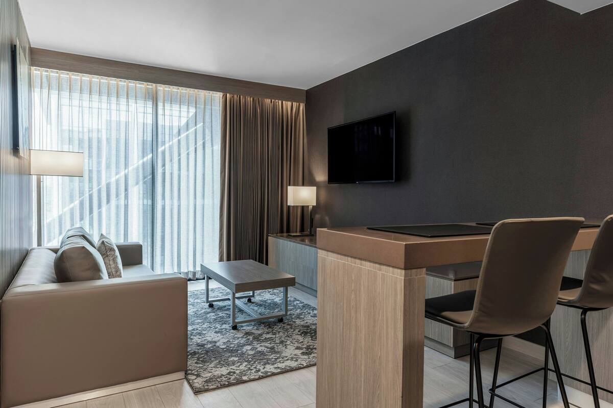 Een moderne hotelkamer met een bank, een klein tafeltje, een bureau met stoelen, een aan de muur bevestigde televisie en grote ramen met gordijnen.