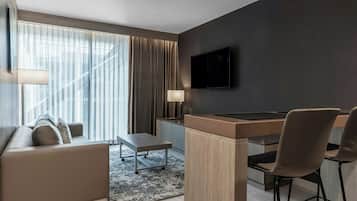 A modern hotel room with a sofa, a small table, a desk with chairs, a TV mounted on the wall, and large windows with curtains.