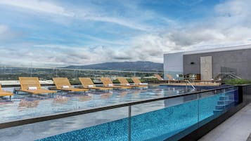 A rooftop infinity pool with wooden loungers, a glass railing, and a view of distant mountains.