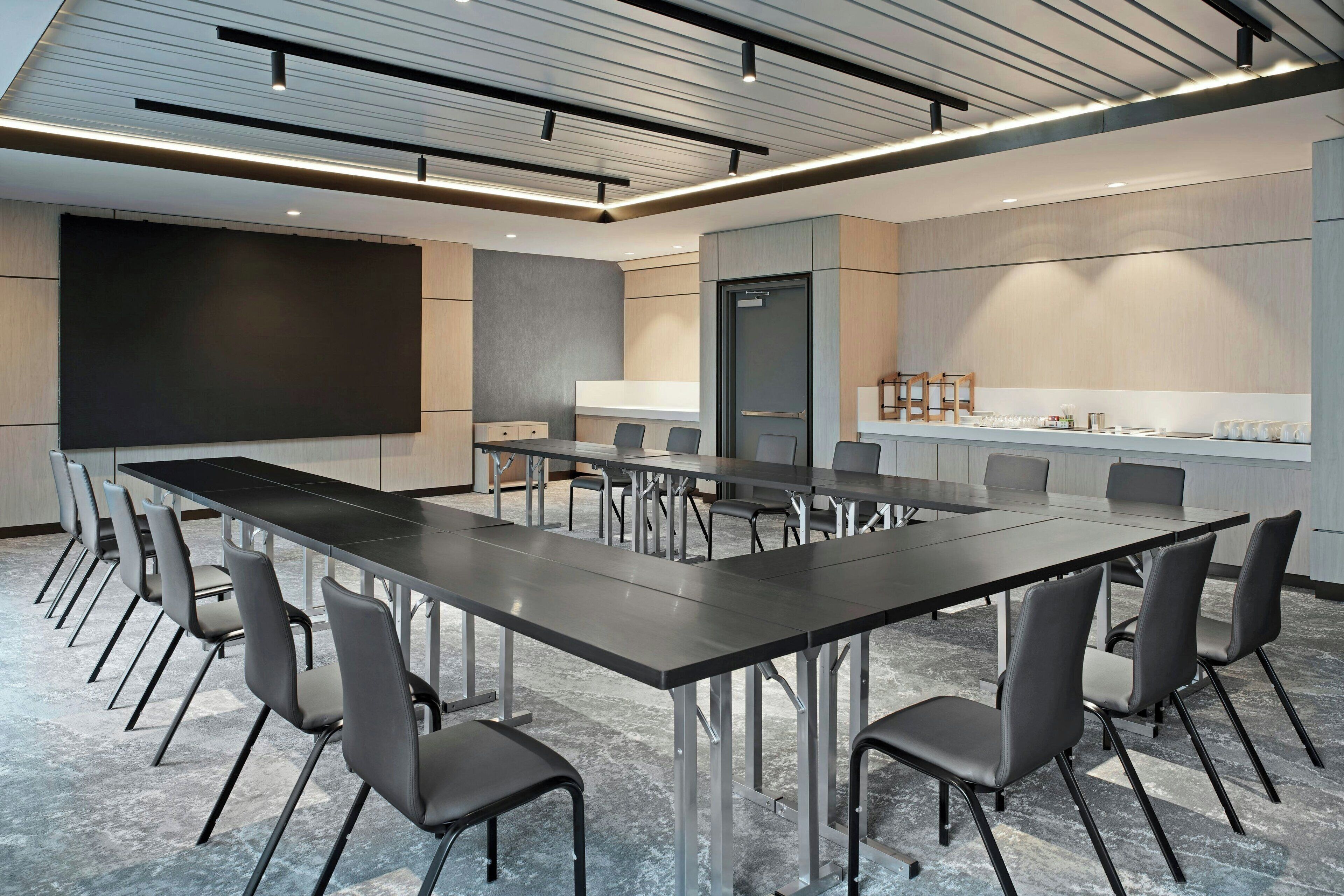 A conference room with a long table, chairs, a projector screen, and a kitchen area.
