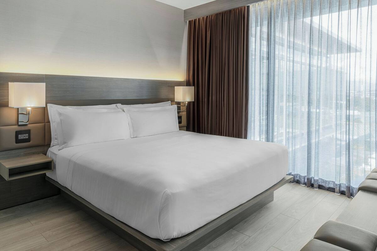 Een moderne hotelkamer met een groot bed, twee nachtkastjes met lampen, uitzicht op de stad door dunne gordijnen, en een houten hoofdbord.