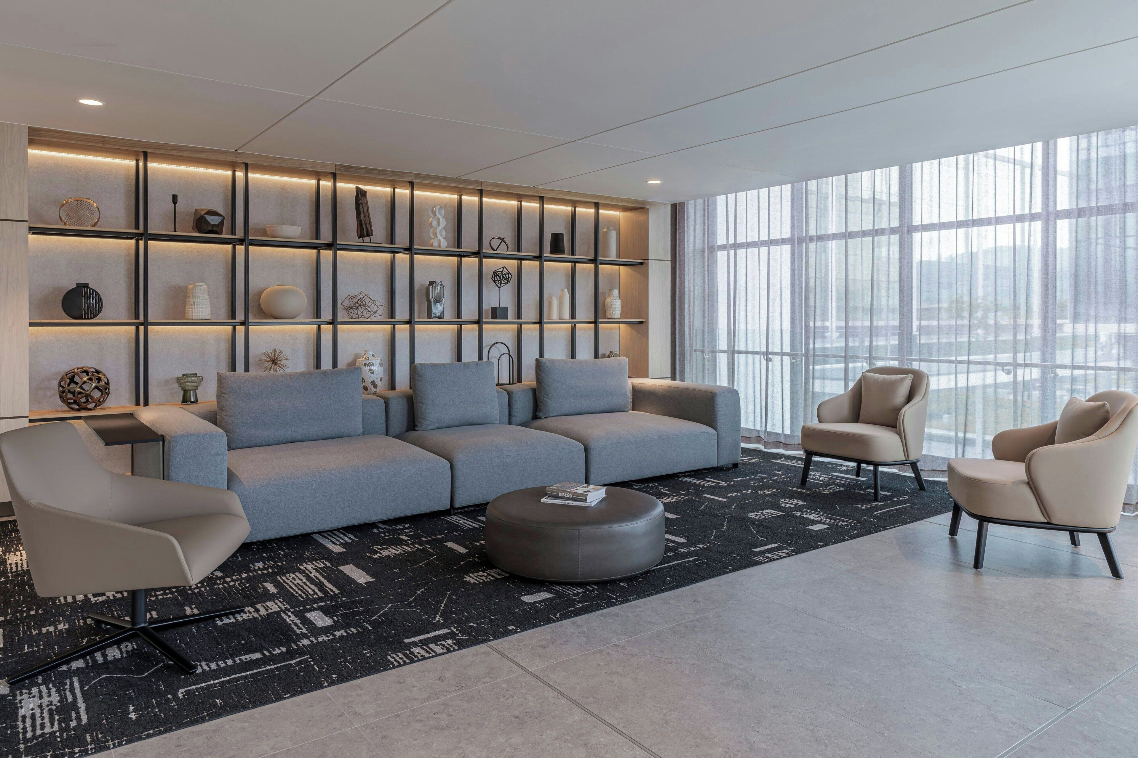 Ein modernes Wohnzimmer mit einem grauen Sofa, zwei Sesseln, einem Couchtisch und einer Wand mit Regalen, auf denen verschiedene dekorative Gegenstände präsentiert werden.