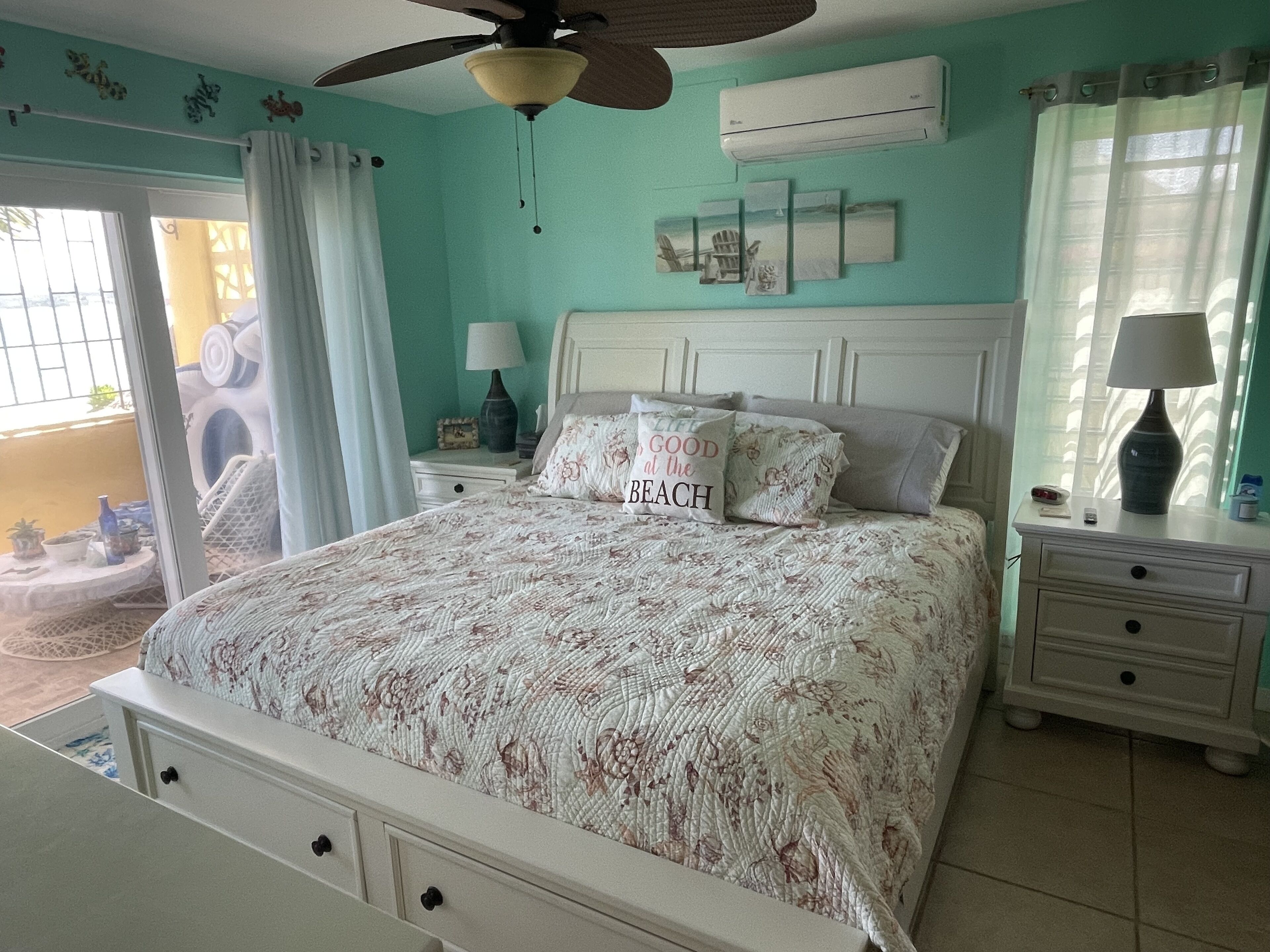 Un dormitorio con cama, mesita de noche, ventilador de techo, aire acondicionado y ventana con cortinas.