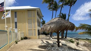 Un bâtiment situé au bord de la plage, doté d’un parasol en chaume, de chaises longues et offrant une vue sur l’océan.