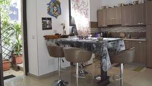 Un espace repas avec une table recouverte d’une nappe à motifs, quatre tabourets de bar, un élément décoratif mural et un coin cuisine doté de meubles en bois et d’un dosseret carrelé.