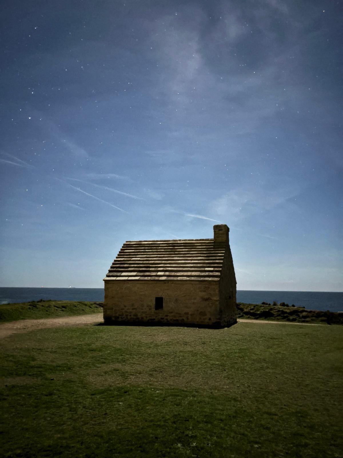 Un pequeño edificio de piedra con techo inclinado y chimenea, ubicado en un área verde con vista despejada al océano y un cielo estrellado.