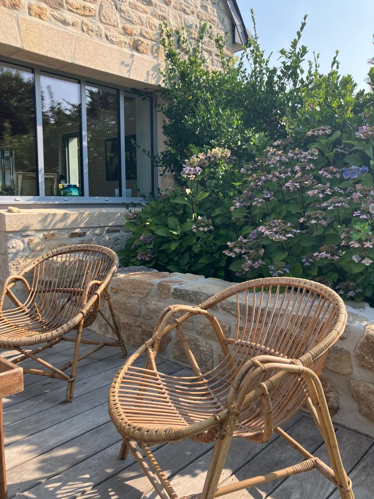 Wicker chairs on a wooden deck, a stone wall, and a bush with pink flowers.