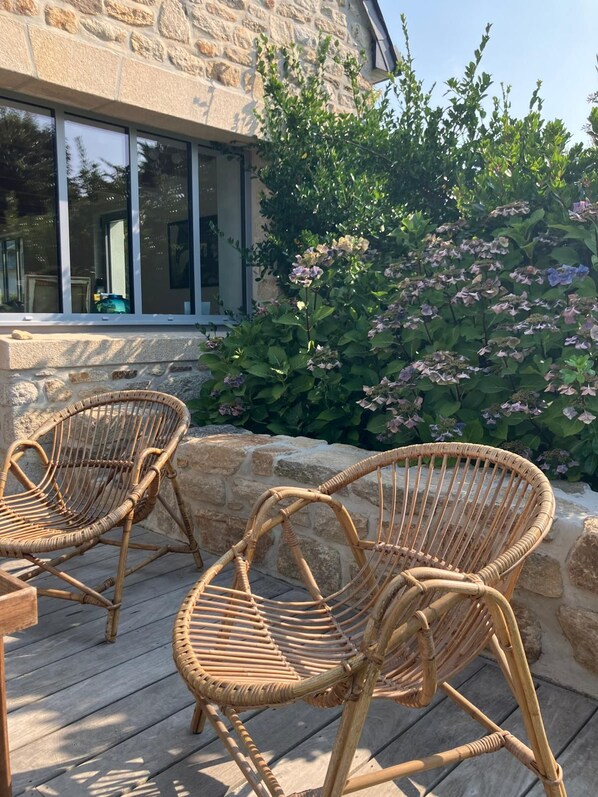 Wicker chairs on a wooden deck, a stone wall, and a bush with pink flowers.