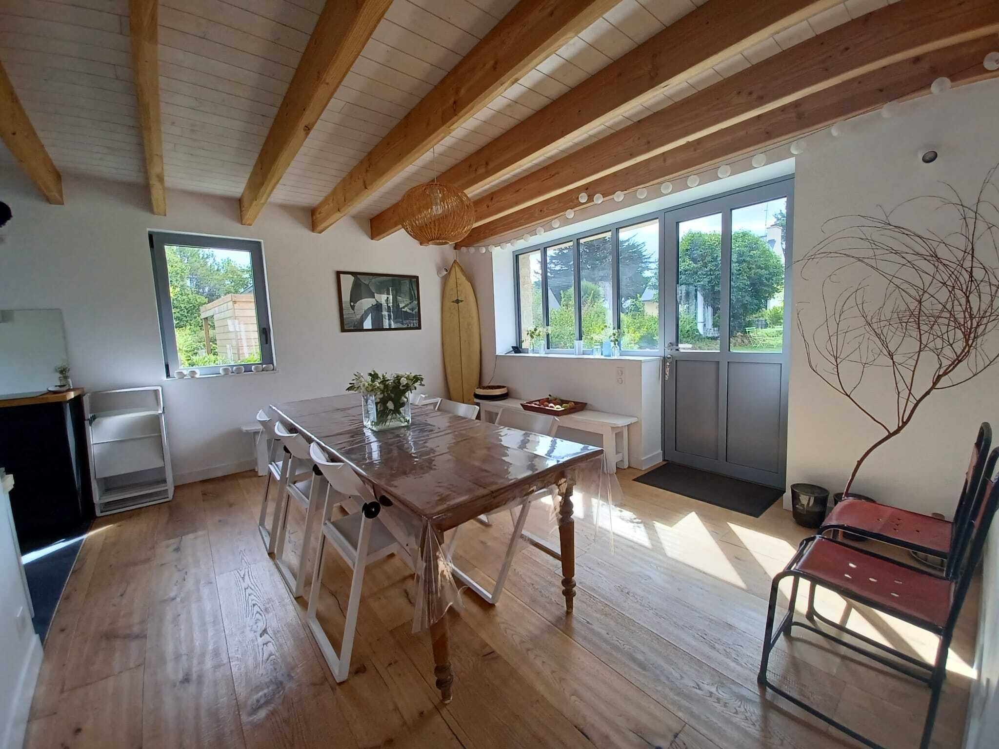 Ein Zimmer mit Holzbalken, einem Esstisch, Stühlen, einer Bank, einem Surfbrett und einem großen Fenster mit Blick auf Grünfläche.