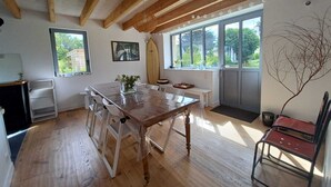 A room with wooden beams, a dining table, chairs, a bench, a surfboard, and a large window with a view of greenery. - beautiful new house near the sea, 3mn walking to the beach, very quiet (Combrit)