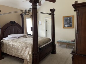 A four-poster bed with a canopy, a bedside table, a chair, and a painting on the wall.