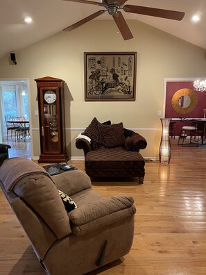 A living room with a ceiling fan, a clock, a couch, a chair, and a dining area with a chandelier.