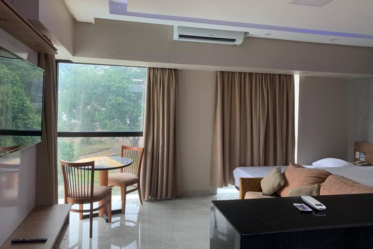 A modern hotel room with a bed, a dining area with a table and chairs, a TV, and a large window with curtains.