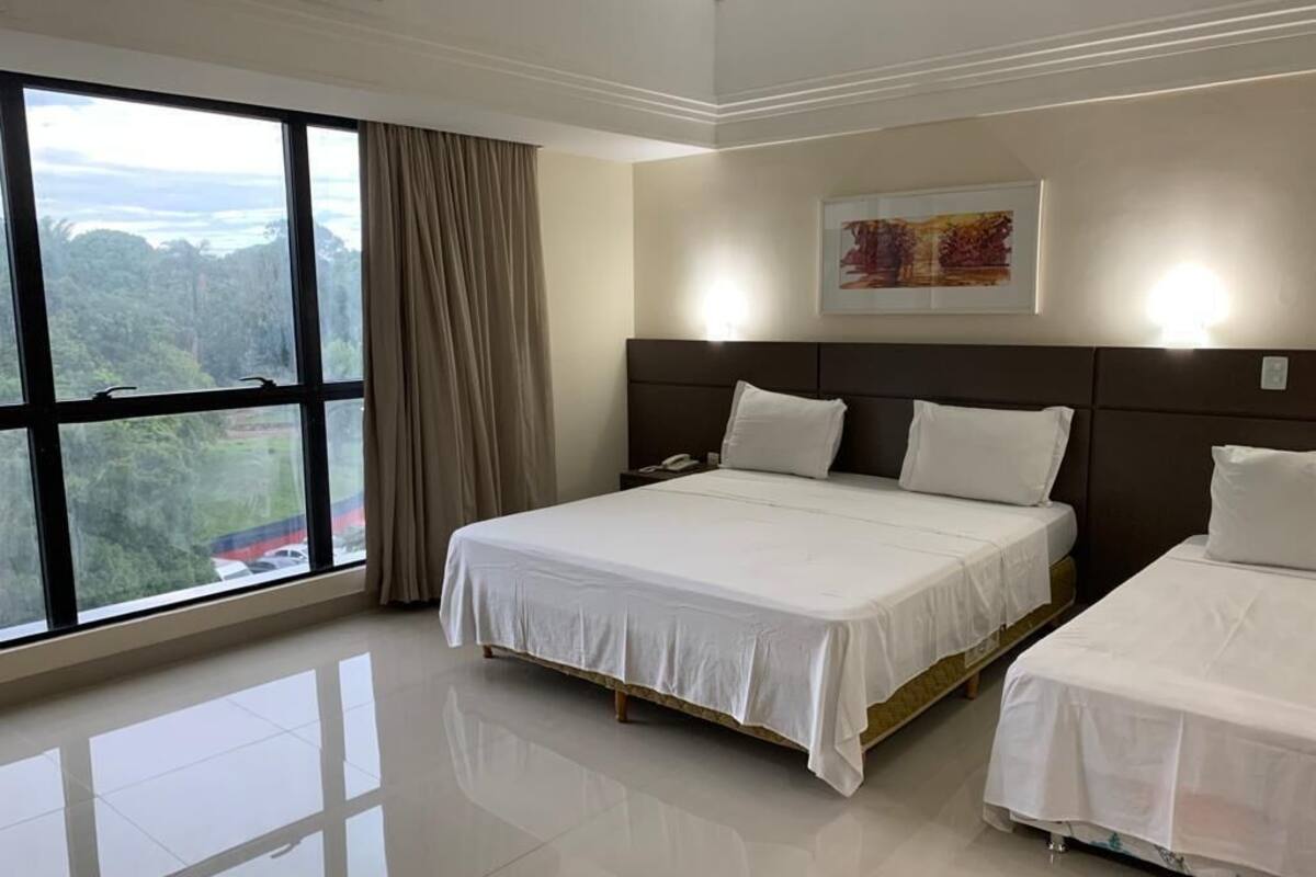 A hotel room with a large window, a bed, a bedside table, and a wall-mounted light.