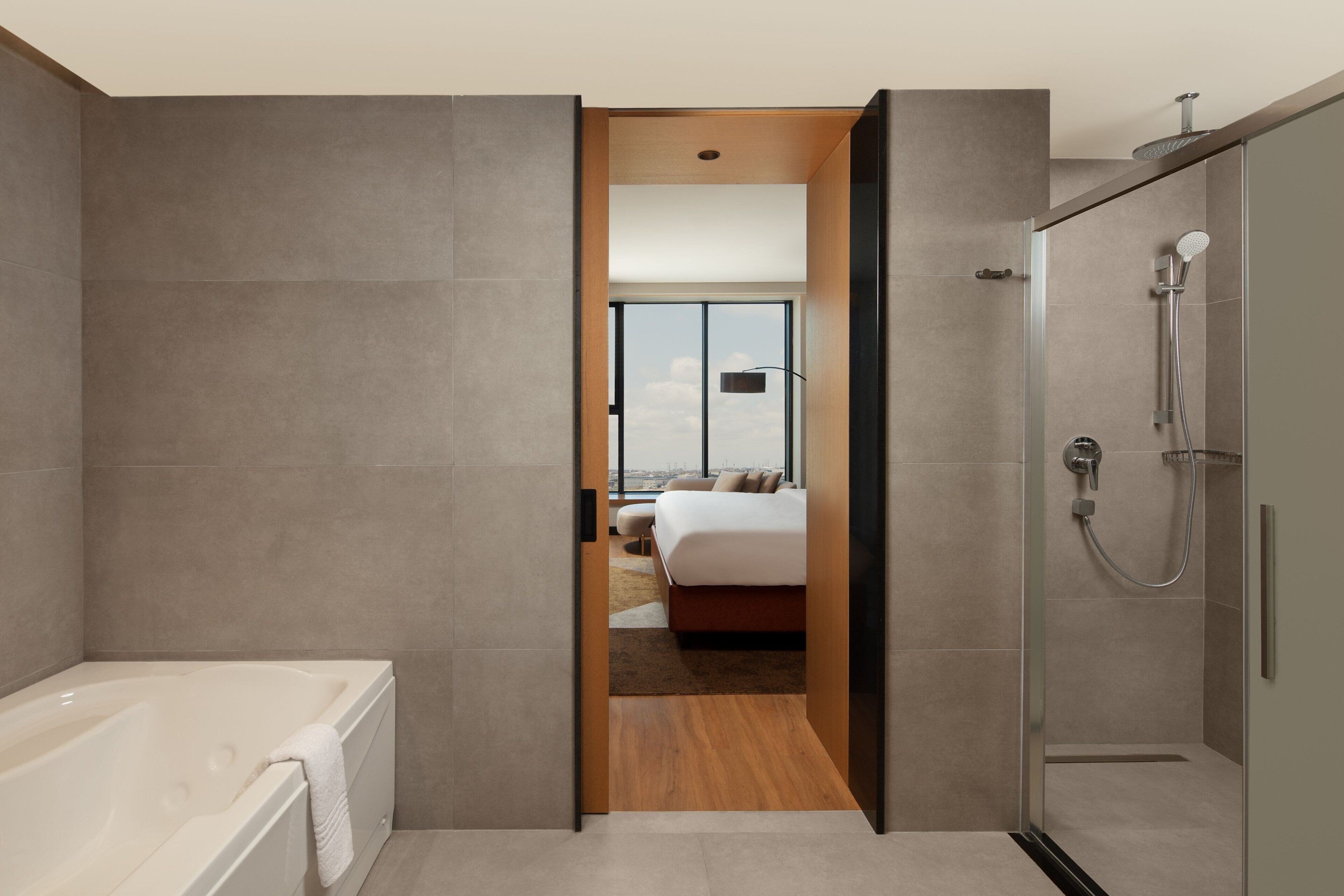 A modern bathroom with a bathtub, shower, and a view of a bedroom with a bed and a cityscape.