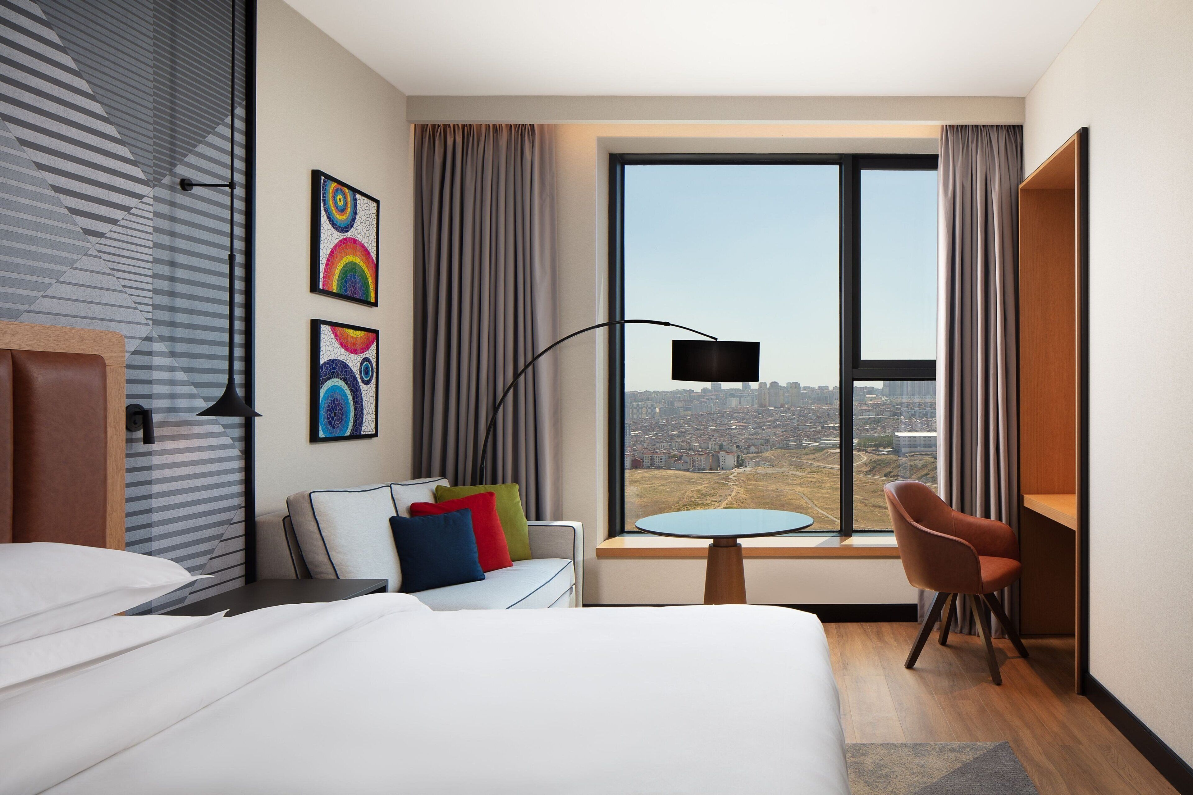 Ett modernt hotellrum med en stor säng, ett skrivbord med en stol, ett runt bord och utsikt över staden genom ett stort fönster.