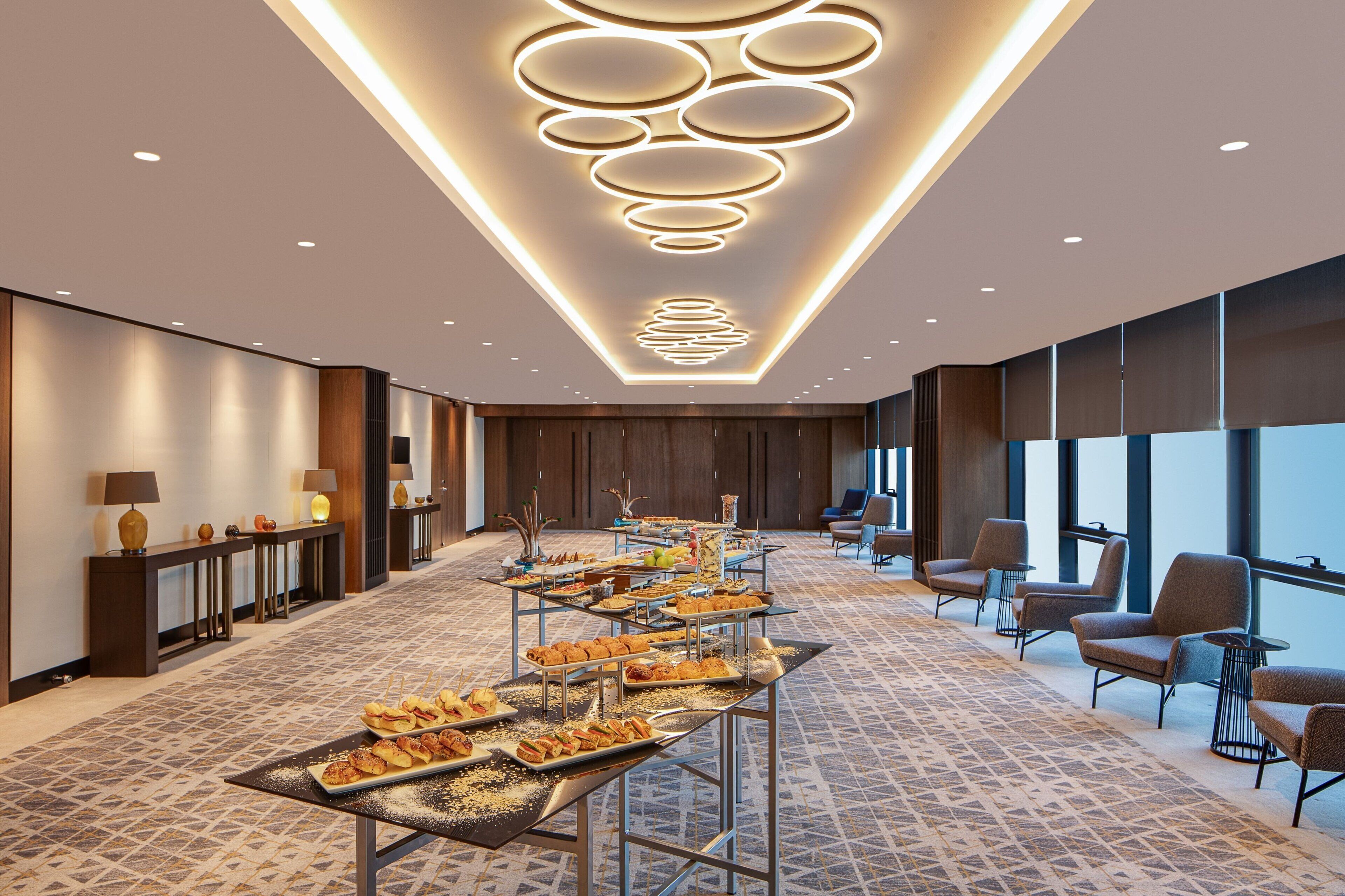 A modern hotel lobby with a long buffet table, comfortable seating, and a decorative ceiling light fixture.