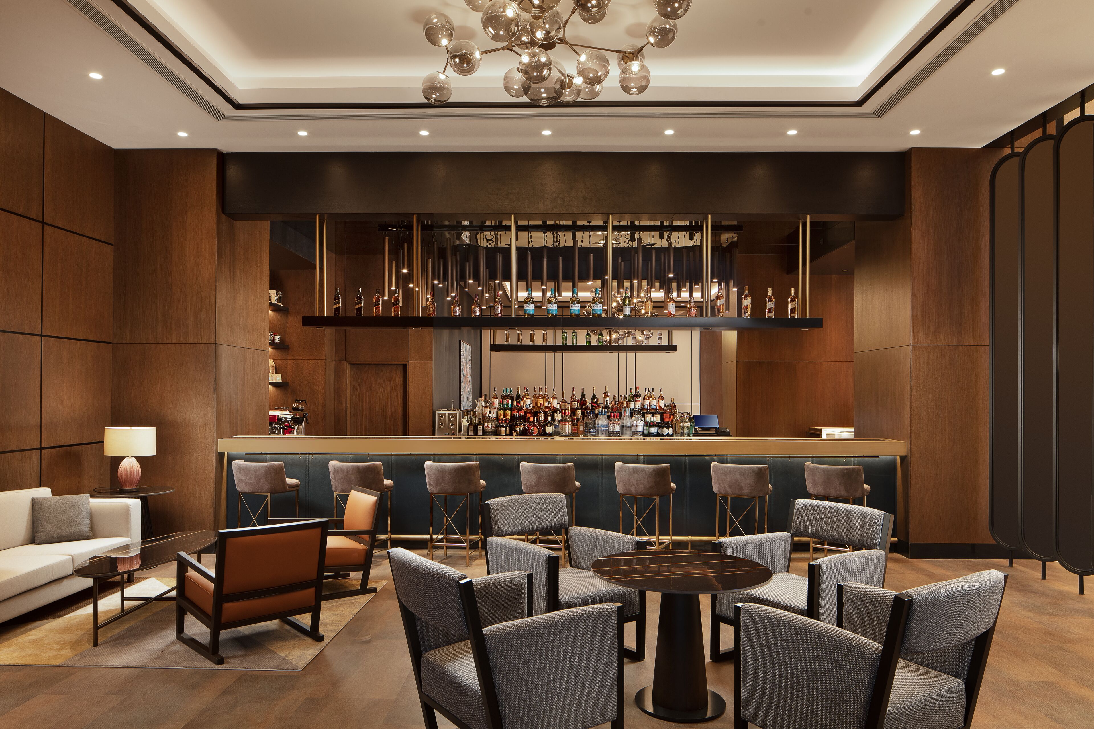 Zona de bar moderna con una barra larga, diversas opciones de asiento, incluyendo taburetes y sillones, y una variedad de botellas exhibidas en estanterías.