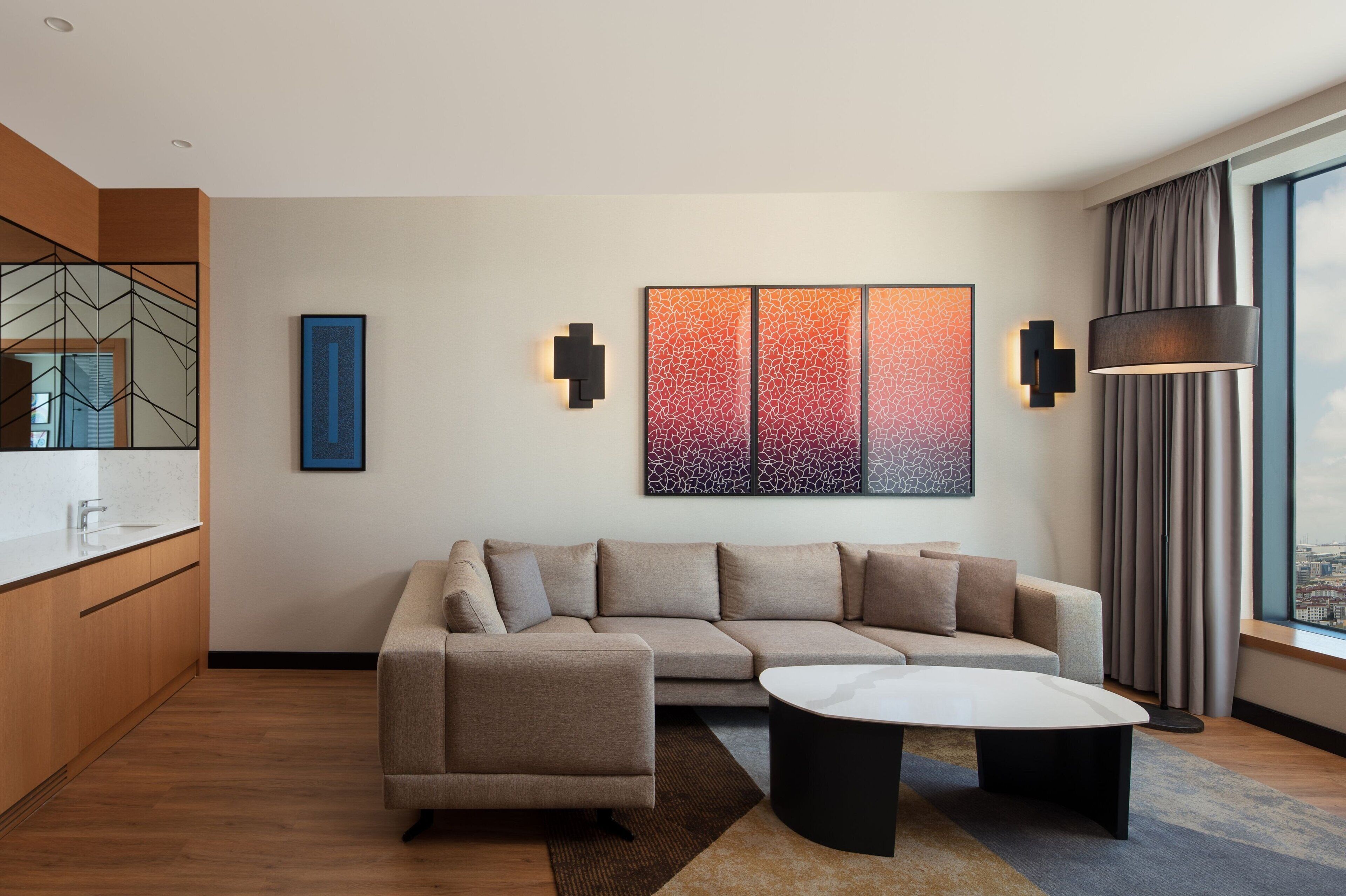 Ein modernes Hotelzimmer mit Ecksofa, rundem Couchtisch, Blick auf die Stadt und Kunstwerken an den Wänden.