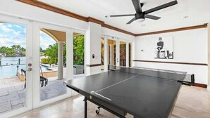A spacious indoor area with a ping pong table, a ceiling fan, and a view of a pool and greenery outside.