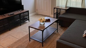 Una sala de estar con una televisión de pantalla plana, una mesa de centro con un florero, un sofá y una ventana con cortinas.