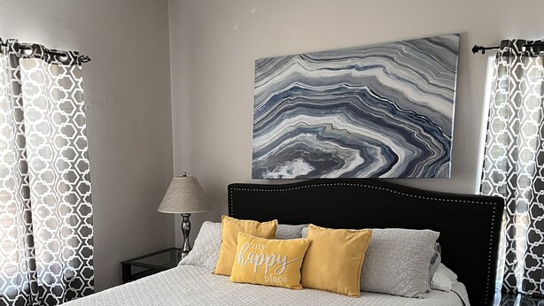 Un dormitorio con una cama, un ventilador de techo, una mesita de noche, una lámpara y un cuadro abstracto grande en la pared.
