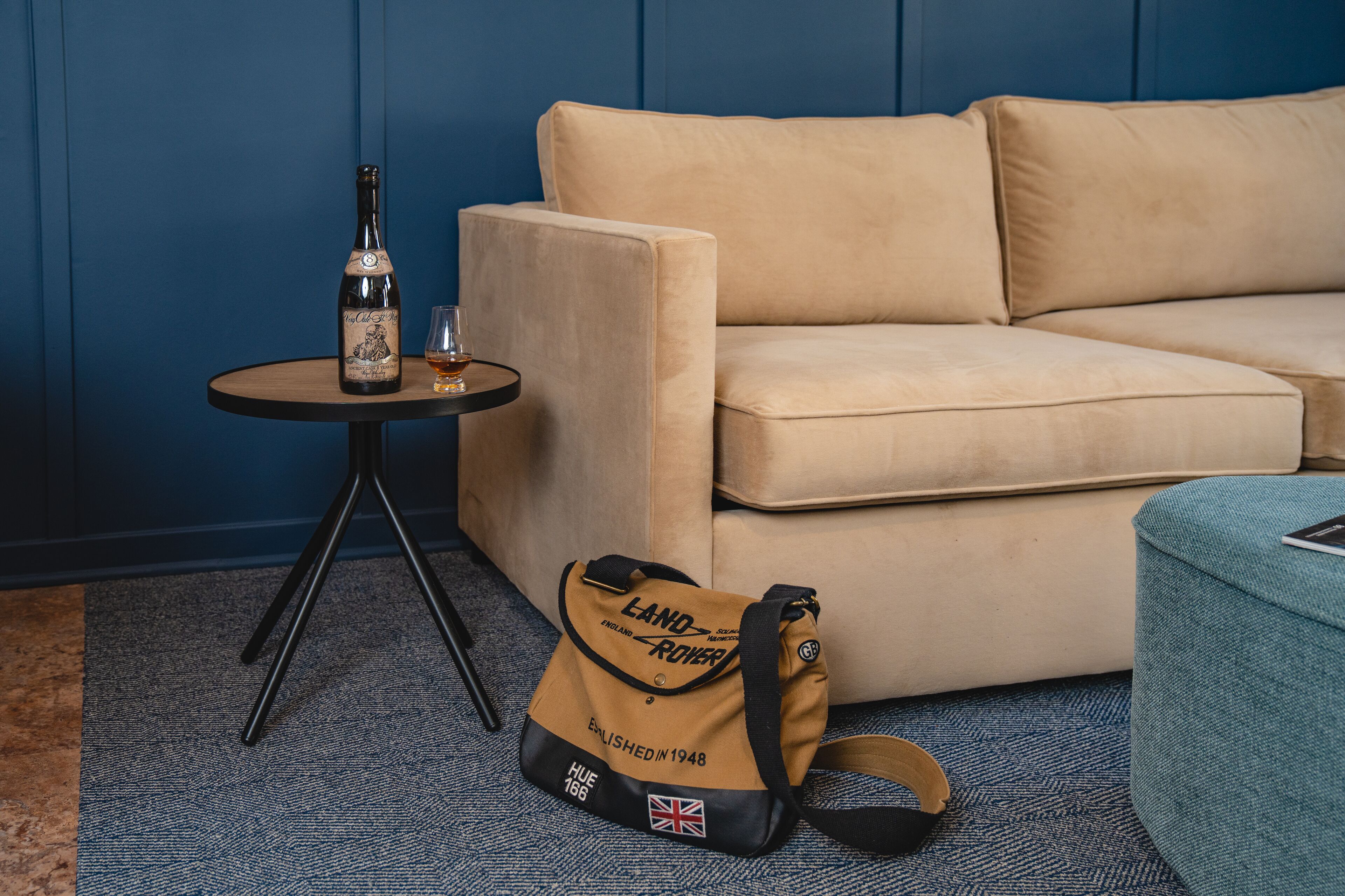 Un sofá color beige, una mesita redonda con una botella y un vaso, y un bolso con el logo de Land Rover sobre una alfombra.