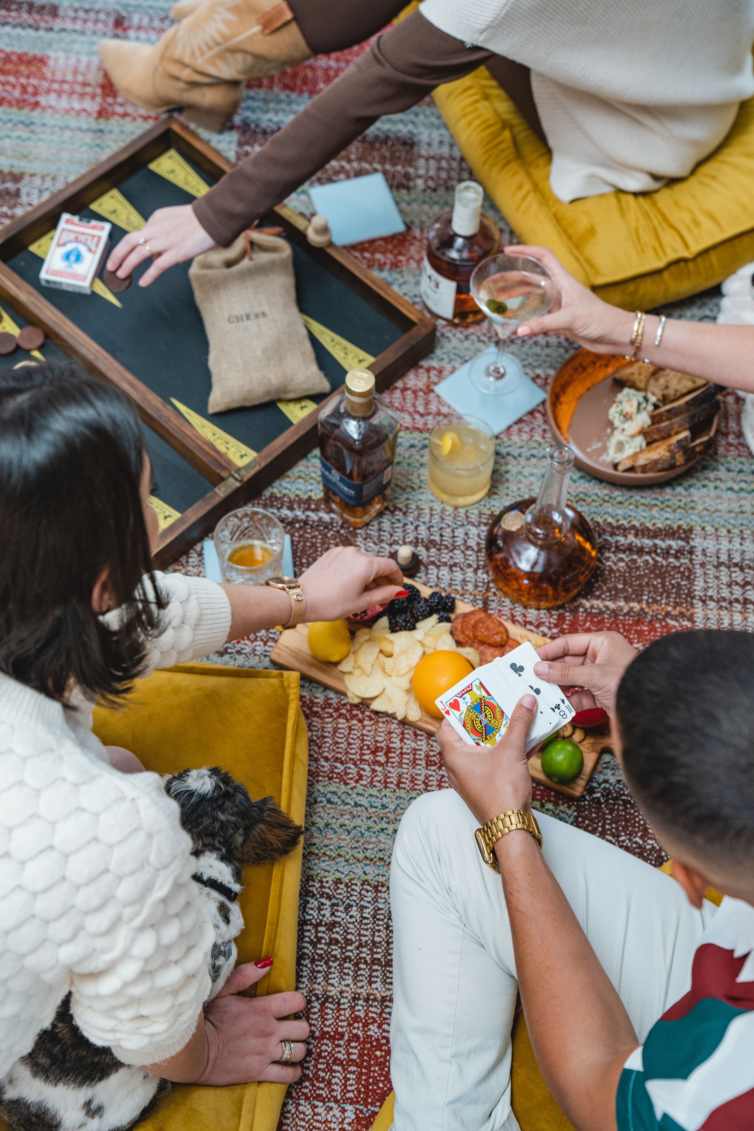A group of people playing cards on a patterned rug with snacks, drinks, and a chessboard.
