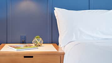 A neatly made bed with white sheets, a wooden nightstand with a book and a decorative plant, and a wall-mounted light fixture.