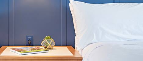 A neatly made bed with white sheets, a wooden nightstand with a book and a decorative plant, and a wall-mounted light fixture.
