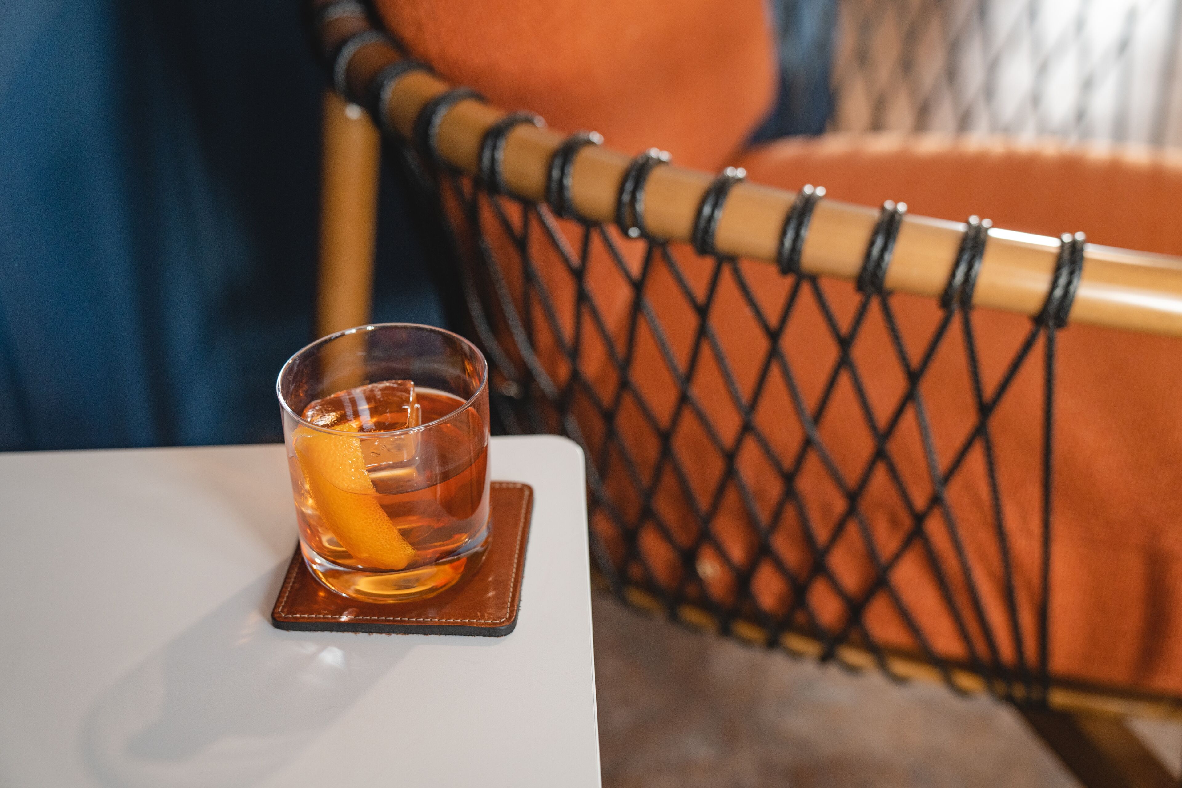 A glass of amber liquid with an orange slice on a coaster, placed on a white table.