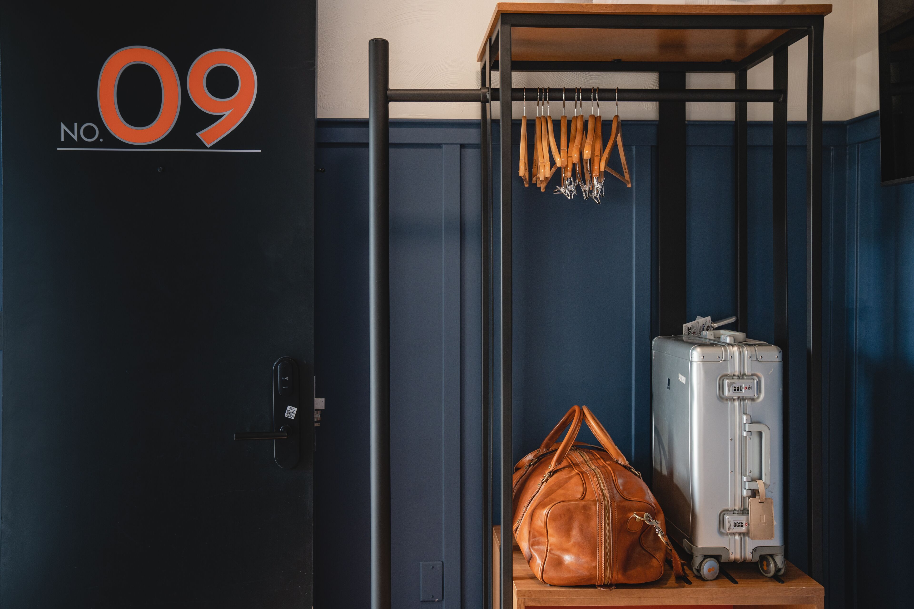 A room with a dark blue door, a wooden shelf with a suitcase and a leather bag, and a wall-mounted rack with tools.