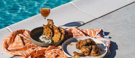 A dining table with a plate of fried chicken and a bowl of salad, set on a beach with a pool in the background.