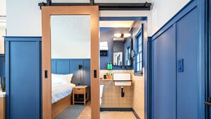 A hotel room with a wooden door leading to a bathroom. The room has a bed, a nightstand, and a small table. The bathroom has a sink, a mirror, and a window. - Bardstown Motor Lodge (Bardstown)