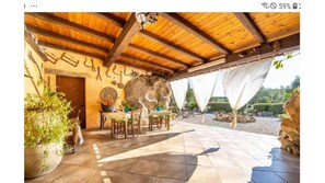 A covered patio area with a stone floor, wooden ceiling beams, a table with chairs, and a large rock wall.