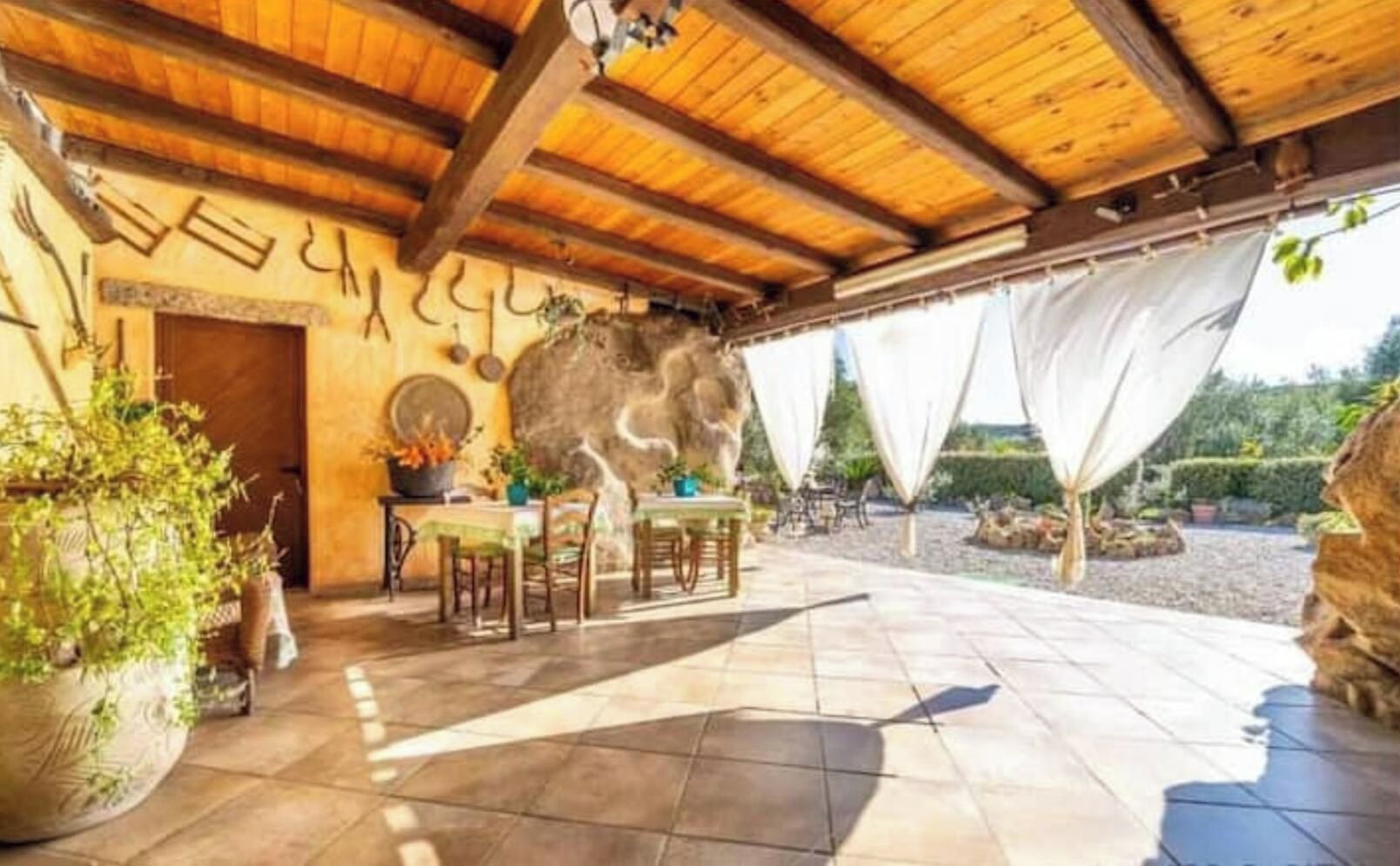 Un espacio al aire libre techado con mesas y sillas, un muro de piedra y plantas en macetas.