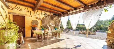Un espacio al aire libre techado con mesas y sillas, un muro de piedra y plantas en macetas.