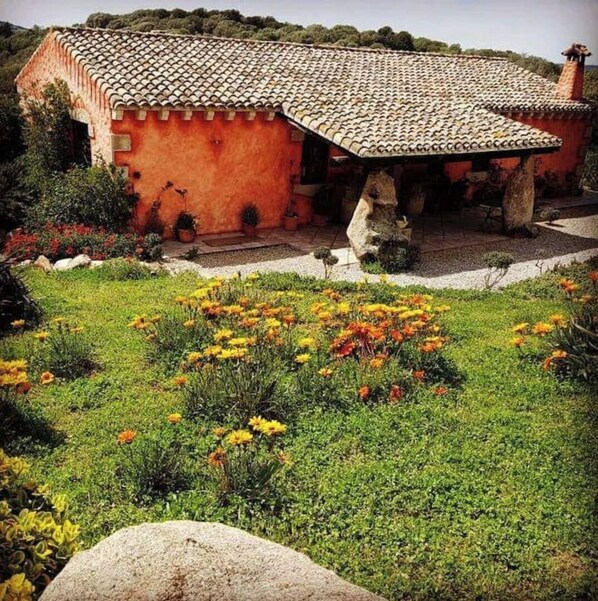 A rustic house with a tiled roof, surrounded by a garden with various flowers and plants.