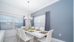 Un coin repas avec une table blanche dressée pour quatre personnes, un lustre et une vue dégagée grâce à de grandes fenêtres équipées de stores.