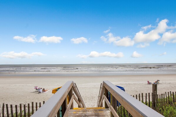 A wooden boardwalk leading to a sandy beach with a clear sky and ocean in the background.