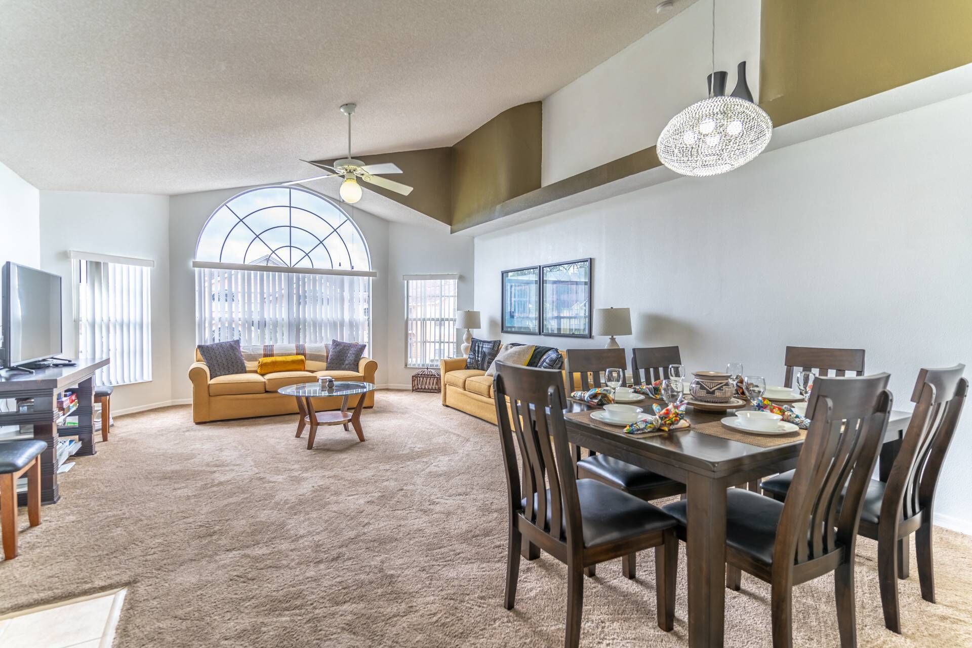 A spacious living room with a large window, a dining area with a table set for four, and a ceiling fan.