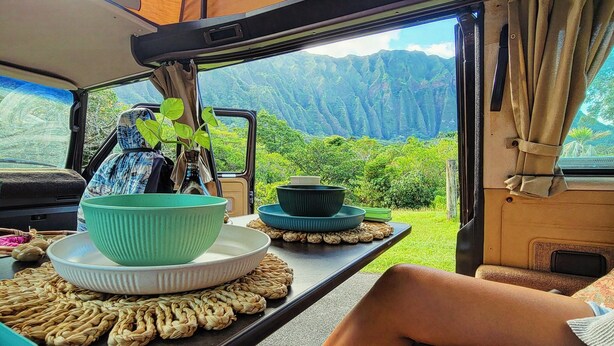 Interior de una campervan con una mesa de comedor preparada para dos personas, una planta en maceta y vistas de montañas a través de la puerta abierta.