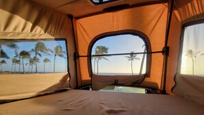 Una habitación con carpa, una cama, una ventana con vistas a palmeras y al mar, y una vista de la playa a través de la ventana.