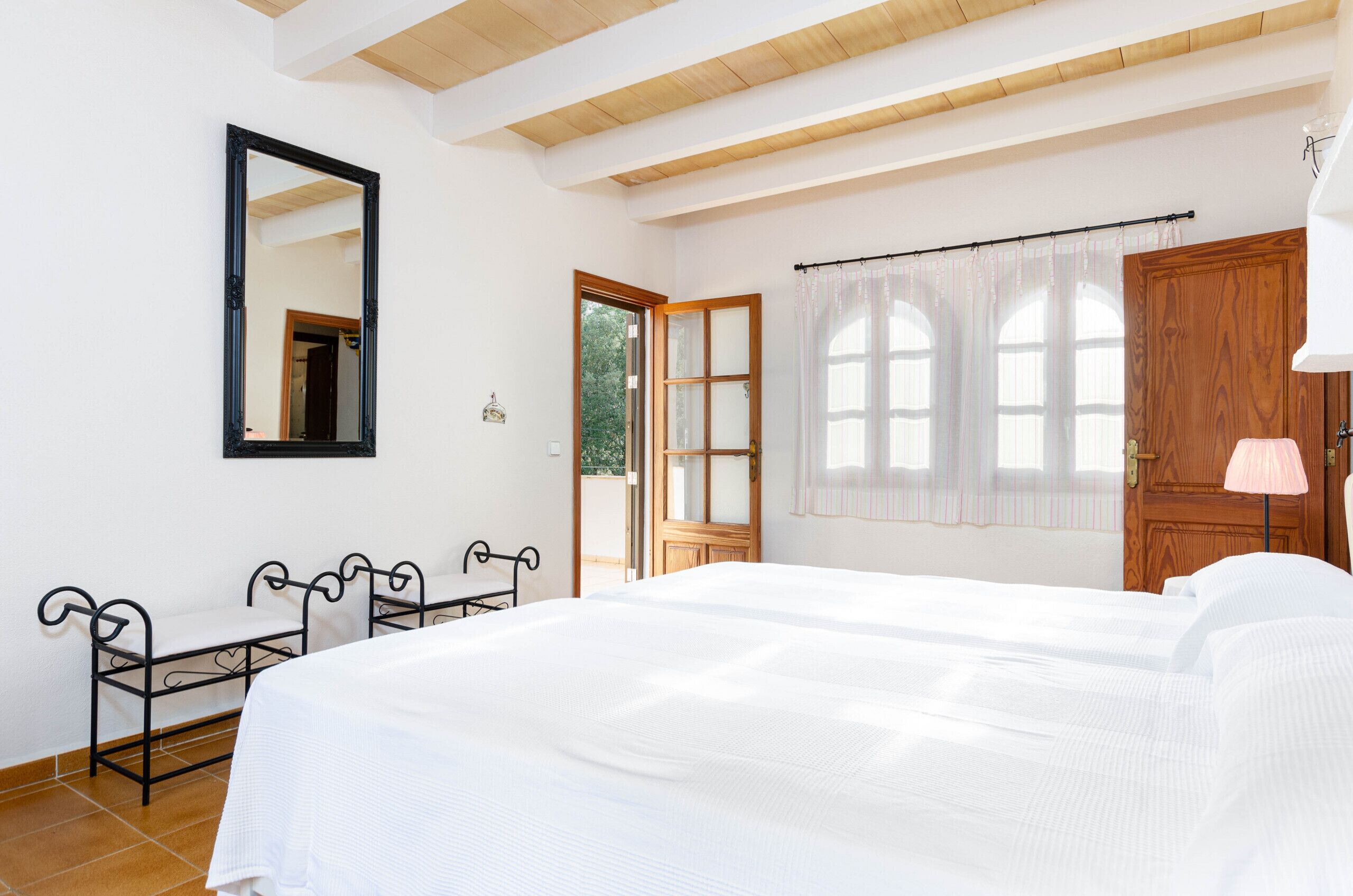 A bedroom with a large bed, a wooden door, a window with curtains, a mirror on the wall, and a small table with chairs.
