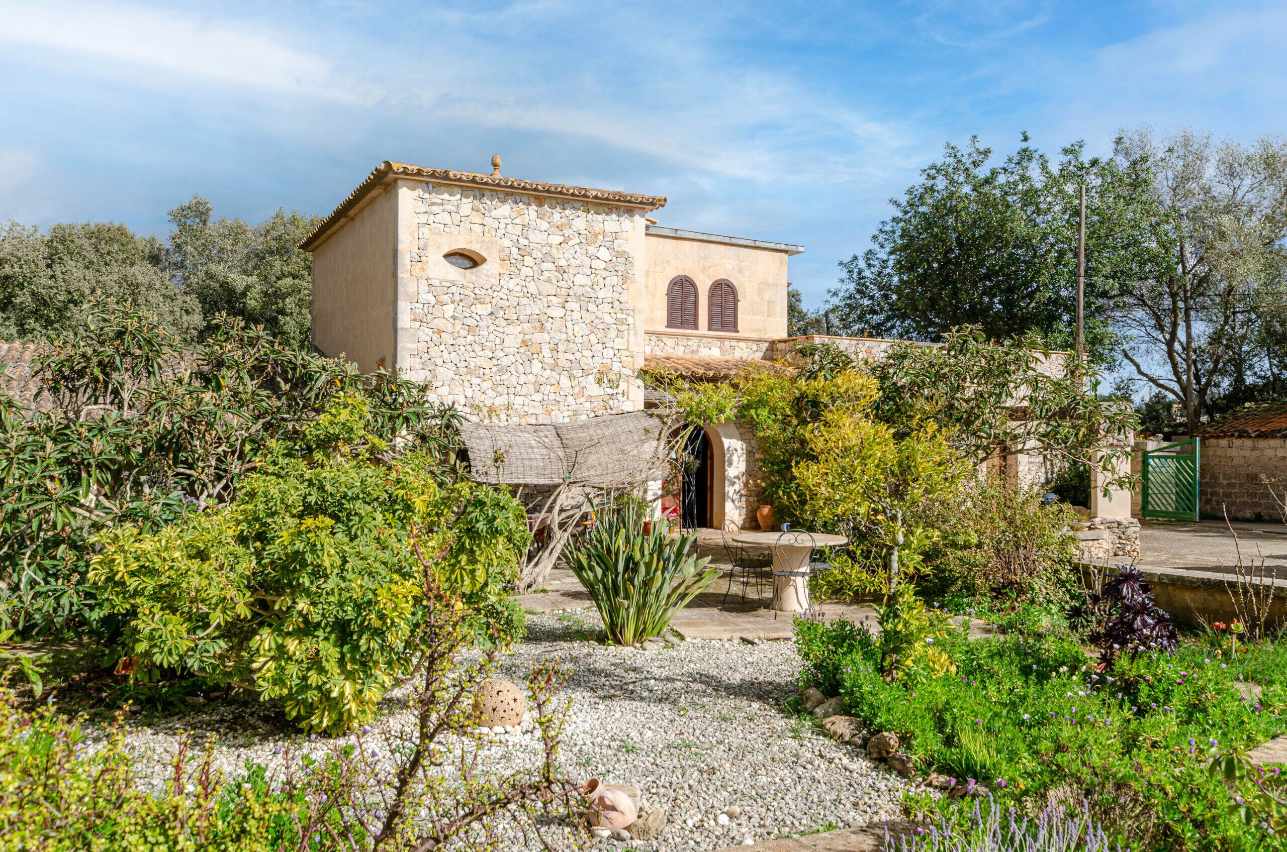 A stone building with a tiled roof, surrounded by a garden with various plants and a gravel path.