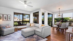 A modern living room with a grey sectional sofa, a coffee table, and a dining area with a wooden table and chairs. Large windows provide views of palm trees and buildings.