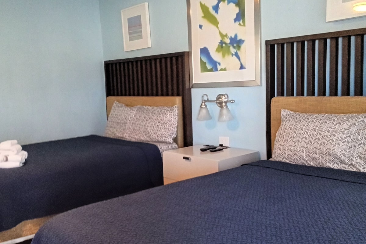 A hotel room with two beds, each with a headboard, a bedside table with a lamp, and a framed picture on the wall.