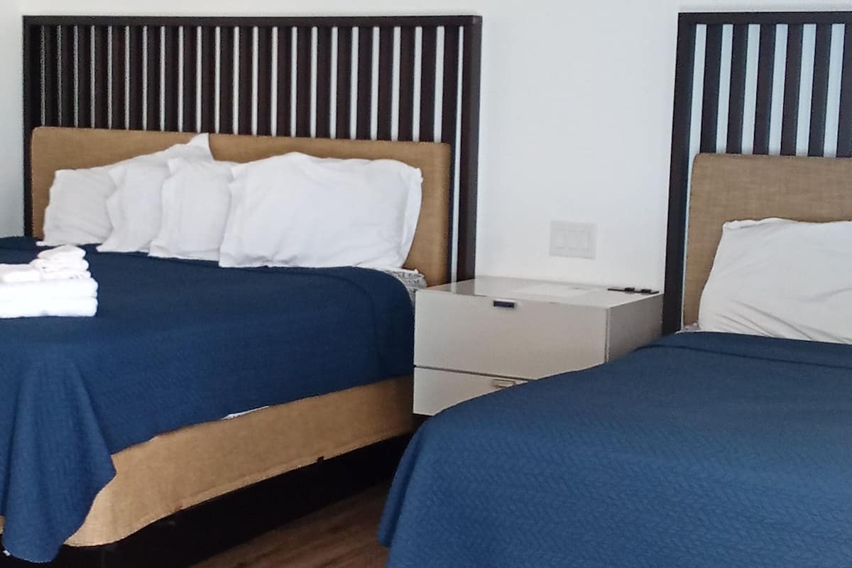 A hotel room with two beds, each with a wooden headboard and blue bedding. There is a white bedside table between the beds, and the room has wooden flooring.